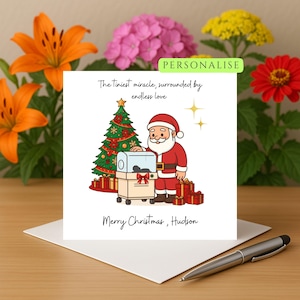 Puede incluir: Tarjeta de Navidad con una ilustración de dibujos animados de Papá Noel, un árbol de Navidad y una incubadora. La tarjeta incluye el texto "El milagro más pequeño, rodeado de amor infinito" y "Feliz Navidad, Hudson".