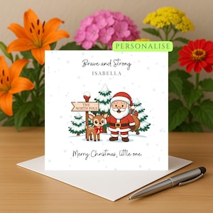 Puede incluir: Tarjeta de Navidad con una ilustración de Papá Noel y un reno. El texto de la tarjeta incluye "Brave and Strong", "Isabella" y "Merry Christmas, little one". El letrero dice "The North Pole". La tarjeta está sobre un sobre blanco.