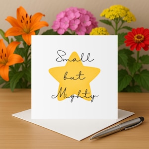 Puede incluir: Una tarjeta blanca con la frase "Small but Mighty" en escritura negra, sobre una estrella amarilla. La tarjeta está sobre un sobre blanco, con un bolígrafo plateado sobre la mesa. Lirios naranjas, hortensias rosas, flores amarillas y una zinnia roja en el fondo.