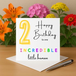 Puede incluir: Una tarjeta de cumpleaños blanca con el número 2 en amarillo, las palabras "Happy Birthday to one" e "Incredible little human" en varios colores. La tarjeta está sobre un sobre blanco con un bolígrafo y rodeada de flores.