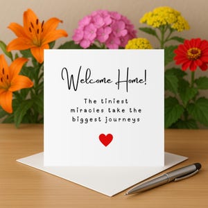 Puede incluir: Una tarjeta de felicitación blanca con las palabras "Welcome Home!" en escritura negra, junto con el mensaje "Los milagros más pequeños hacen los viajes más grandes" y un corazón rojo. La tarjeta está sobre un sobre blanco, con un bolígrafo plateado. Flores naranjas, rosas, rojas y amarillas están en el fondo.