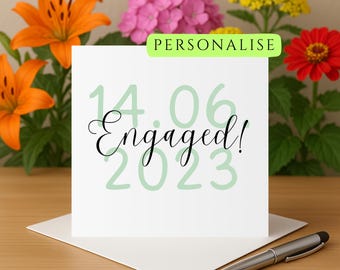 Könnte beinhalten: Eine weiße Karte mit dem Text "14.06 Verlobt! 2023" in grüner und schwarzer Schrift. Die Karte liegt auf einer Holzoberfläche mit Blumen im Hintergrund.