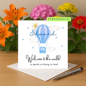 Puede incluir: Una tarjeta de felicitación con una ilustración de globo aerostático azul y blanco, el nombre "Edward" en escritura cursiva y el texto "Welcome to the world". La tarjeta está sobre un sobre blanco, con un bolígrafo encima. Flores en el fondo.