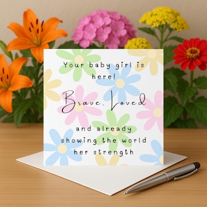 Puede incluir: Una tarjeta de felicitación blanca con ilustraciones de flores en colores pastel y el texto "Your baby girl is here! Brave. Loved and already showing the world her strength." Un bolígrafo plateado descansa sobre la tarjeta. Flores coloridas en el fondo.