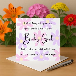 Puede incluir: Una tarjeta de felicitación con el texto "Thinking of you as you welcome your Baby Girl into the world with so much love and courage". La tarjeta está decorada con flores rosas y moradas y está sobre un sobre blanco. Lirios naranjas y otras flores están en el fondo.