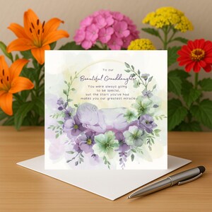 Puede incluir: Una tarjeta de felicitación con el texto "To our Beautiful Granddaughter" y una ilustración en acuarela de un bebé durmiendo rodeado de flores moradas y verdes. Un bolígrafo plateado descansa sobre la tarjeta.