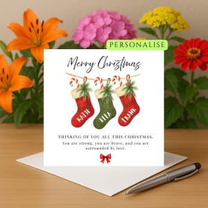 Puede incluir: Tarjeta de Navidad con la frase "Merry Christmas" y tres calcetines navideños con los nombres "Katie", "Ella" y "Frank". También incluye el texto "Thinking of you all this Christmas".