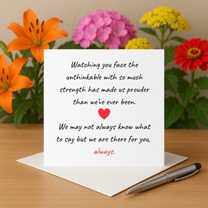 Può includere: Un biglietto di auguri bianco con il testo "Watching you face the unthinkable with so much strength has made us prouder than we've ever been." Un cuore rosso è al centro. Una penna d'argento è appoggiata sulla carta. Fiori sullo sfondo.