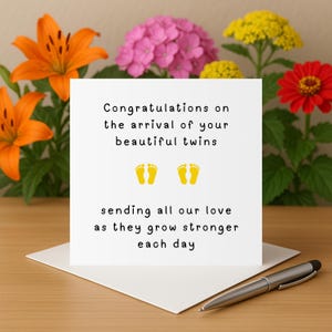 Puede incluir: Una tarjeta blanca con el texto "Felicitaciones por la llegada de tus hermosos gemelos" y huellas amarillas. La tarjeta está sobre una superficie de madera, con un bolígrafo y flores en el fondo.