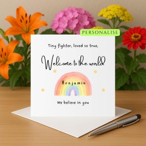Puede incluir: Tarjeta de felicitación blanca con la frase "Welcome to the world" y un diseño de arcoíris. La tarjeta incluye el nombre "Benjamin" y las frases "Tiny fighter, loved so true" y "We believe in you". Flores y un bolígrafo en el fondo.