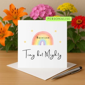 Puede incluir: Una tarjeta blanca con un diseño de arcoíris en acuarela y el nombre "Harrison" en el centro. Las palabras "Tiny but Mighty" están escritas debajo. La tarjeta está sobre un sobre blanco, con un bolígrafo al lado. Flores en el fondo.