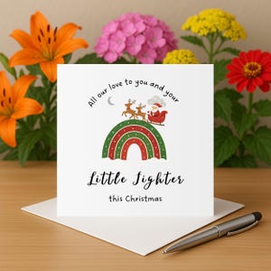 Puede incluir: Una tarjeta de felicitación blanca con el texto "All our love to you and your Little Fighter this Christmas." La tarjeta presenta un diseño de arcoíris con Papá Noel en un trineo tirado por renos. La tarjeta está sobre un sobre blanco.