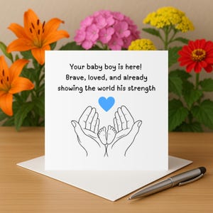 Puede incluir: Una tarjeta blanca con el texto "Your baby boy is here! Brave, loved, and already showing the world his strength." La tarjeta presenta un corazón azul y una ilustración de manos acunando pies de bebé. Un bolígrafo plateado descansa sobre la mesa.