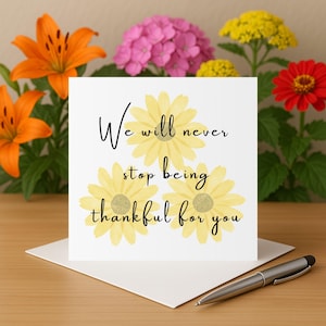 Puede incluir: Una tarjeta de felicitación blanca con ilustraciones de margaritas amarillas y el texto "We will never stop being thankful for you." La tarjeta está sobre una superficie de madera con un bolígrafo y rodeada de flores.