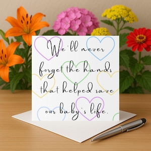 Puede incluir: Una tarjeta blanca con el texto "We'll never forget the hands that helped save our baby's life." está decorada con formas de corazón pastel. Un bolígrafo plateado descansa sobre el sobre. El fondo presenta lirios naranjas, hortensias rosas y flores amarillas y rojas.