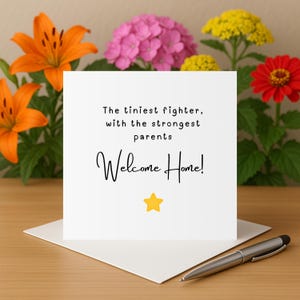 Puede incluir: Una tarjeta de felicitación blanca con el texto "The tiniest fighter, with the strongest parents, Welcome Home!" y una estrella amarilla. La tarjeta está sobre un sobre blanco, con un bolígrafo plateado. Lirios naranjas, flores rosas y amarillas en el fondo.