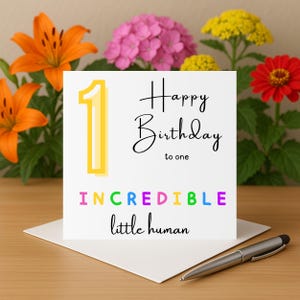 Puede incluir: Una tarjeta de cumpleaños blanca con el texto "Happy Birthday to one" y "Incredible little human" en letras coloridas. Un número 1 amarillo está a la izquierda. La tarjeta está sobre un sobre blanco con un bolígrafo. Las flores están en el fondo.