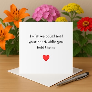 Puede incluir: Una tarjeta de felicitación blanca con el texto "I wish we could hold your heart while you hold theirs" y un corazón rojo. La tarjeta está sobre un sobre blanco, con un bolígrafo plateado sobre la mesa. Lirios naranjas, flores rosas y amarillas en el fondo.
