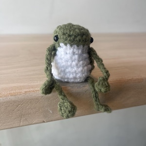 May include: A handmade crochet frog in shades of green and white. The frog has black bead eyes and is sitting with its legs extended. The frog is made of yarn and has a textured appearance. The frog is a cute and whimsical item.