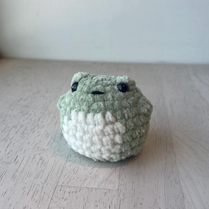 May include: A handmade crochet frog in shades of green and white. The plush toy has black safety eyes and a small, stitched mouth. The frog is a soft, rounded shape, perfect for a child's toy or nursery decor.