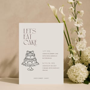 以下が含まれることがあります： 「LET'S EAT CAKE」の文字とケーキのイラストが描かれたクリーム色のサイン。スイスメレンゲバタークリームを使ったレモンブルーベリーケーキと、ヘーゼルナッツプラリネを使ったチョコレートシーソルトキャラメルのケーキのフレーバーが記載されています。背景には白い花があります。