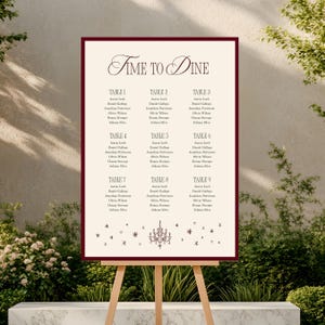 Peut inclure: Un plan de table de mariage crème avec une bordure bordeaux, présenté sur un chevalet en bois. Le tableau présente le texte "Time to Dine" dans une écriture élégante, ainsi que les noms des invités organisés par numéro de table.