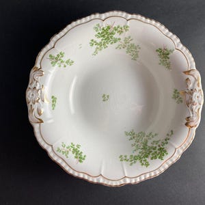 May include: A white ceramic serving bowl with a gold rim and green floral design. The bowl has two handles on the sides.