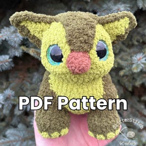 Puede incluir: Un peluche hecho a mano en tonos verdes y marrones, con ojos grandes y expresivos y una nariz rosa. El texto "PDF Pattern" se muestra en la parte delantera. El juguete tiene un aspecto suave y texturizado, lo que sugiere una construcci&oacute;n de ganchillo o tejido.
