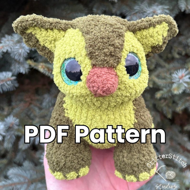 Puede incluir: Un peluche hecho a mano en tonos verdes y marrones, con ojos grandes y expresivos y una nariz rosa. El texto "PDF Pattern" se muestra en la parte delantera. El juguete tiene un aspecto suave y texturizado, lo que sugiere una construcci&oacute;n de ganchillo o tejido.