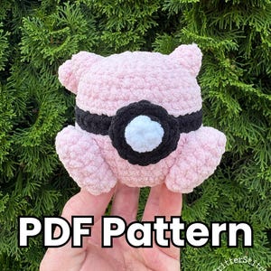 May include: A light pink crocheted plush toy featuring a black band around its center with a white circular accent in the middle. The text PDF Pattern is displayed in white letters with a black outline at the bottom of the image.