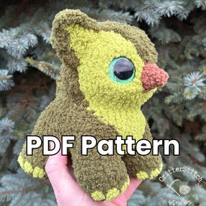 Puede incluir: Un peluche con forma de aguacate, con grandes ojos azules y una nariz marr&oacute;n. El juguete est&aacute; hecho de tela texturizada verde y marr&oacute;n. El texto "PDF Pattern" est&aacute; visible. El juguete se sostiene en una mano.