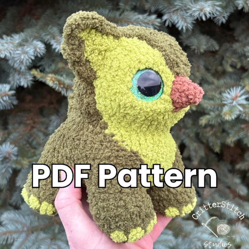 Puede incluir: Un peluche con forma de aguacate, con grandes ojos azules y una nariz marr&oacute;n. El juguete est&aacute; hecho de tela texturizada verde y marr&oacute;n. El texto "PDF Pattern" est&aacute; visible. El juguete se sostiene en una mano.