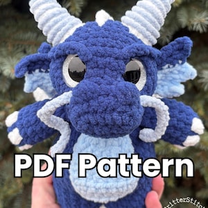 以下が含まれることがあります： 手作りの青いかぎ針編みのドラゴン。水色と白のアクセントが施されています。ドラゴンは大きく光沢のある目を持ち、フレンドリーな表情をしています。「PDF Pattern」の文字がドラゴンの体に表示されています。