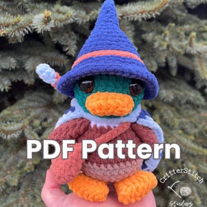 May include: Crochet pattern for a plush duck wearing a blue wizard hat with a red band. The duck has a teal body, orange beak, and brown and orange feet. The pattern is available as a PDF download. The text "PDF Pattern" is in large white letters on a red background. The text "CritterStitch Studios" is in white letters on a black background with a stylized needle and yarn.