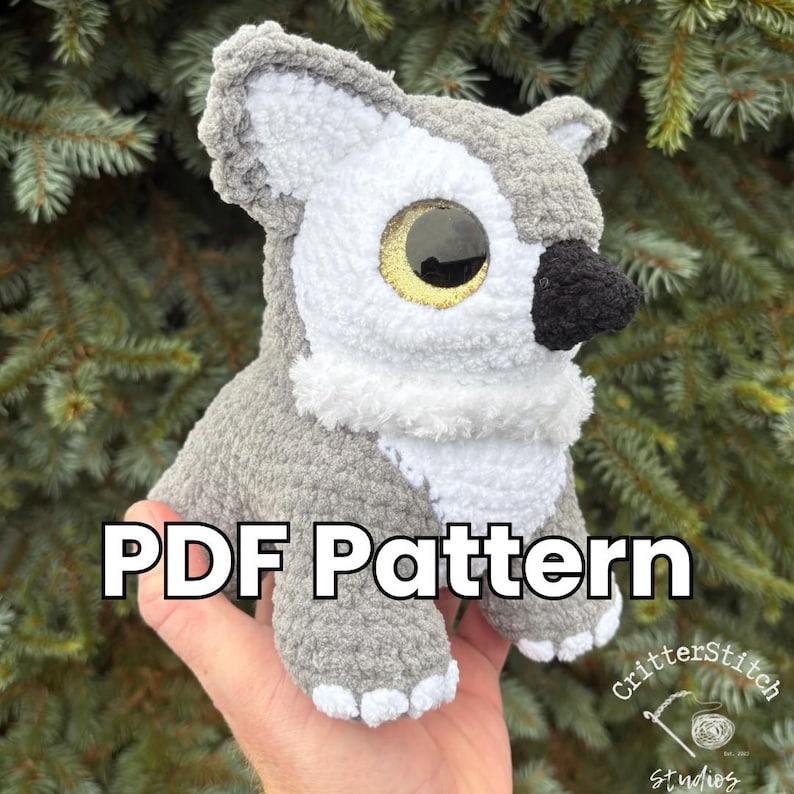 Puede incluir: Un peluche de lobo tejido a ganchillo en gris, blanco y negro, con un detalle de ojo dorado. El texto "PDF Pattern" se muestra en el peluche. El peluche se sostiene en una mano contra un fondo de follaje verde.