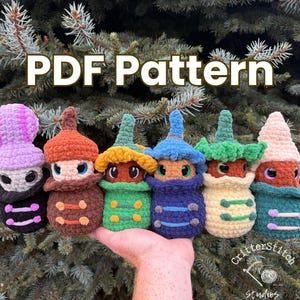 May include: A collection of six colorful, crocheted mushroom-shaped figures. Each figure has a unique color scheme, including purple, orange, green, blue, and cream. The text "PDF Pattern" is displayed at the top. The figures are held in a hand.