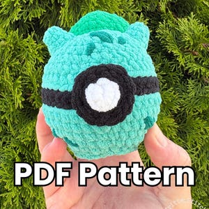 May include: A round, teal, crocheted plush toy featuring a black horizontal band across the centre with a white circular accent in the middle. The surface has a textured, fuzzy appearance. The text PDF Pattern is printed in white with a black outline.