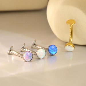 May include: A selection of opal labret studs in shades of purple, white, and blue. One gold-coloured curved barbell stud with a white opal stone is also visible. The jewellery is presented on a white surface.