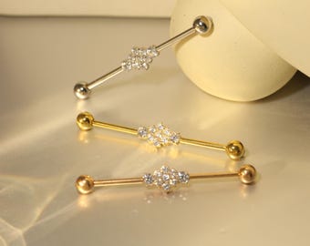 Op de afbeelding: Drie industriële barbells in zilver, goud en roségoud. Elke barbell heeft een cluster van kleine, heldere edelstenen in het midden en een bal aan elk uiteinde. De sieraden worden gepresenteerd op een reflecterend oppervlak.