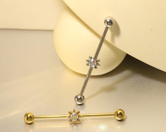 Op de afbeelding: Twee industriële barbell oorbellen. Eén is zilverkleurig, de andere goudkleurig. Elk heeft een stervormig midden met een heldere, iriserende edelsteen. De uiteinden van elke barbell zijn rond. De oorbellen worden tegen een neutrale achtergrond getoond.