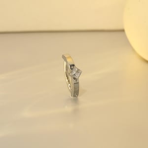 Puede incluir: Un anillo plateado con una gema cuadrada y transparente. El anillo tiene un diseño moderno con una banda ligeramente curvada y un engaste elevado para la gema. El anillo se muestra sobre un fondo neutro.