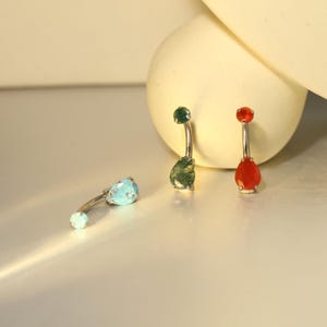 以下が含まれることがあります： カーブしたシルバーバーのベリーボタンリング3つ。1つはターコイズストーン、もう1つはグリーンのストーン、3つ目は赤いストーンです。各リングの上部には小さな丸い石が付いています。宝石は涙の形をしています。