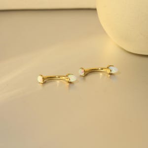 May include: Two gold-colored curved eyebrow piercing jewelry with opal-like gemstones at each end. The jewelry is displayed on a light-colored surface, with a soft, diffused light.
