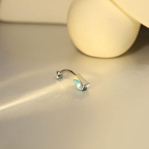 May include: A silver-toned belly button ring with two light blue gemstones. One gemstone is round, and the other is a rectangular shape with a curved bar.