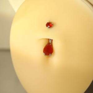 May include: A belly button piercing with a teardrop-shaped red gem pendant and a small, round red gem above. The jewelry is silver-colored and set against a light skin tone.