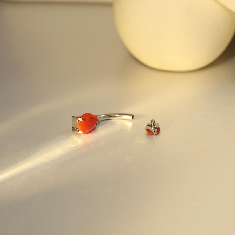 May include: A silver belly button ring with a teardrop-shaped, orange gemstone and a small, matching gem screw-on ball. The jewelry is displayed on a light-colored surface.