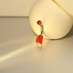 May include: A silver belly button ring with a curved barbell design. The ring features a small, round, red gemstone at the top and a larger, teardrop-shaped, red gemstone at the bottom. The gemstones have a faceted cut.