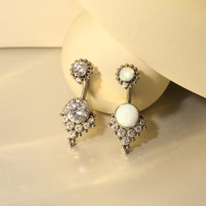 Peut inclure: Deux piercings de nombril argentés. L'un présente une grosse pierre précieuse ronde et transparente en bas et une plus petite en haut. L'autre a une grosse opale blanche en bas et une plus petite en haut.