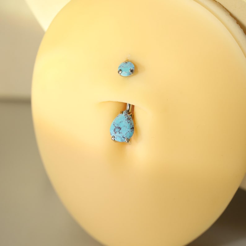May include: A belly button piercing with a teardrop-shaped turquoise stone and a smaller matching stone above. The jewelry is silver-toned and set against a light yellow background.