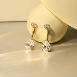 May include: Two belly button rings. One has a gold-colored curved bar and a pear-shaped clear stone. The other has a silver-colored curved bar and a pear-shaped clear stone. Both have a small, round clear stone at the top of the bar.